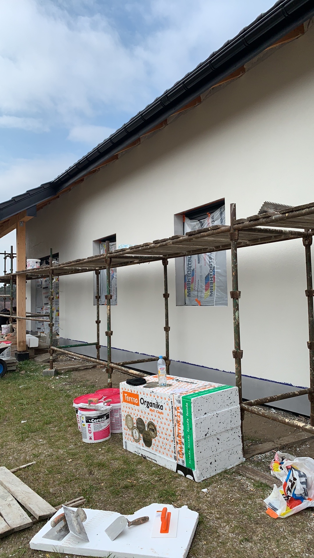 Świeżo otynkowana elewacja budynku jednorodzinnego z zamaskowanymi oknami, rusztowanie, wiadro z farbą Ceresit CT 74, opakowanie styropianu Termo Organika i narzędzia murarskie na trawie.
