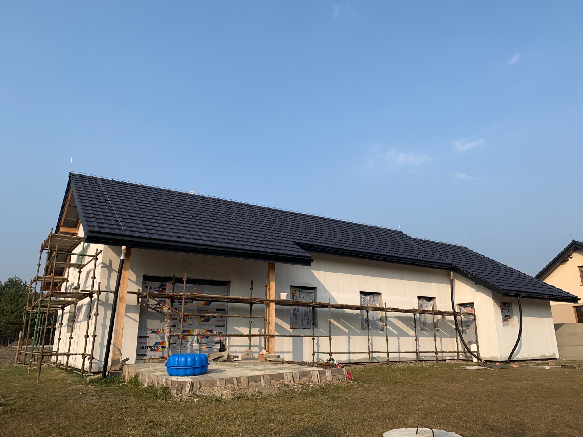 Dom w trakcie prac elewacyjnych: ocieplony, z rusztowaniami, częściowo zabezpieczone okna folią, widoczny niebieski pojemnik i elementy materiałów budowlanych.