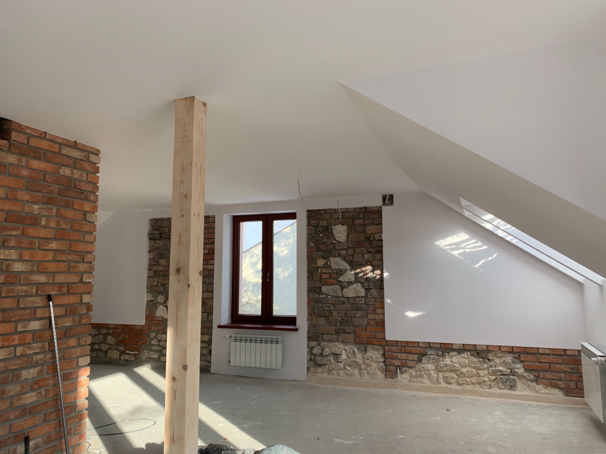 Wnętrze poddasza w trakcie remontu: fragmenty ścian z cegły i kamienia, drewniany słup, świeżo pomalowane na biało ściany i sufit, okno z ciemną ramą, grzejnik.