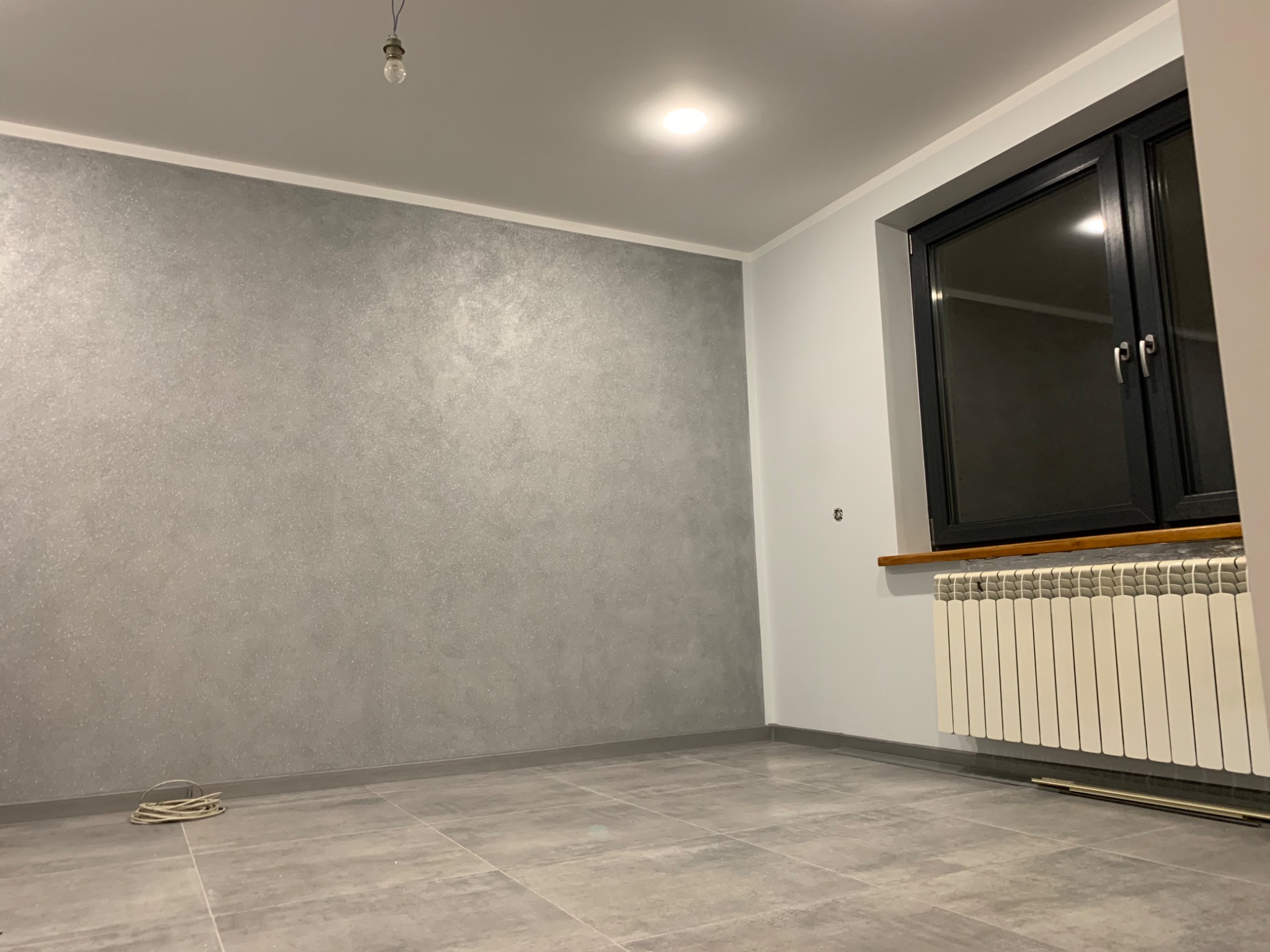 Pokój po wykonaniu gładzi dekoracyjnej w odcieniach szarości, z widocznym grzejnikiem pod oknem i płytkami podłogowymi. Na suficie widoczna żarówka na kablu, sugerująca prace wykończeniowe.