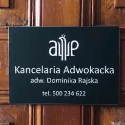 Tabliczka informacyjna Kancelarii Adwokackiej adw. Dominiki Rajskiej z logo na ciemnozielonym tle, zamocowana na drewnianych drzwiach.