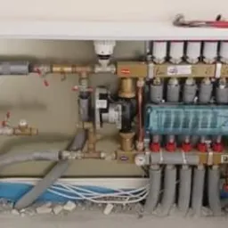 Rozdzielacz ogrzewania podłogowego z izolowanymi rurami, zamontowany w białej szafce wnękowej, widoczny klucz nastawny na górnej krawędzi.