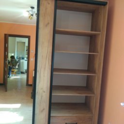 WREM SPÓŁKA Z OGRANICZONĄ ODPOWIEDZIALNOŚCIĄ - Szafa wnękowa z przesuwnymi drzwiami lustrzanymi i drewnianym wykończeniem, widoczne półki i szuflada. W tle pomarańczowa ściana i widok na kuchnię.