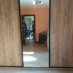 WREM SPÓŁKA Z OGRANICZONĄ ODPOWIEDZIALNOŚCIĄ - Szafa przesuwna z lustrem, drewnopodobne fronty w ciepłym odcieniu. W lustrze odbija się fragment pokoju z widocznym oświetleniem sufitowym i osobami w tle.