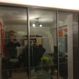 WREM SPÓŁKA Z OGRANICZONĄ ODPOWIEDZIALNOŚCIĄ - Szafa wnękowa z przesuwnymi, szklanymi drzwiami z czarnymi ramami. Widoczne odbicie pokoju z oświetleniem i osobą robiącą zdjęcie.