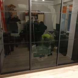 WREM SPÓŁKA Z OGRANICZONĄ ODPOWIEDZIALNOŚCIĄ - Nowoczesna szafa wnękowa z przesuwnymi, grafitowymi drzwiami lustrzanymi, odbijającymi wnętrze pokoju. Widoczne detale wykończenia i odbicie osoby robiącej zdjęcie.