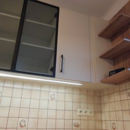 WREM SPÓŁKA Z OGRANICZONĄ ODPOWIEDZIALNOŚCIĄ - Białe szafki kuchenne z czarnymi uchwytami i szklanymi frontami, oświetlenie LED pod szafkami, drewniane półki na ścianie z płyty imitującej drewno.