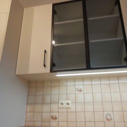 WREM SPÓŁKA Z OGRANICZONĄ ODPOWIEDZIALNOŚCIĄ - Białe szafki kuchenne wiszące z czarnymi uchwytami i szklanymi frontami, oświetlenie podszafkowe, kremowe płytki z motywem filiżanek w tle.