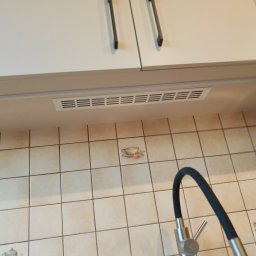 WREM SPÓŁKA Z OGRANICZONĄ ODPOWIEDZIALNOŚCIĄ - Białe szafki kuchenne z czarnymi uchwytami, oświetlenie podszafkowe LED i beżowe płytki z motywem filiżanki, widoczny fragment baterii kuchennej.