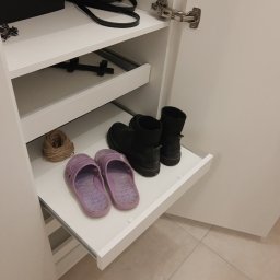 WREM SPÓŁKA Z OGRANICZONĄ ODPOWIEDZIALNOŚCIĄ - Wysuwana półka w szafie z zawartością: klapki, buty i sznurek. Widoczne okucia szafy i fragment podłogi z płytek. Ujęcie z bliska, wnętrze szafy w jasnej tonacji.