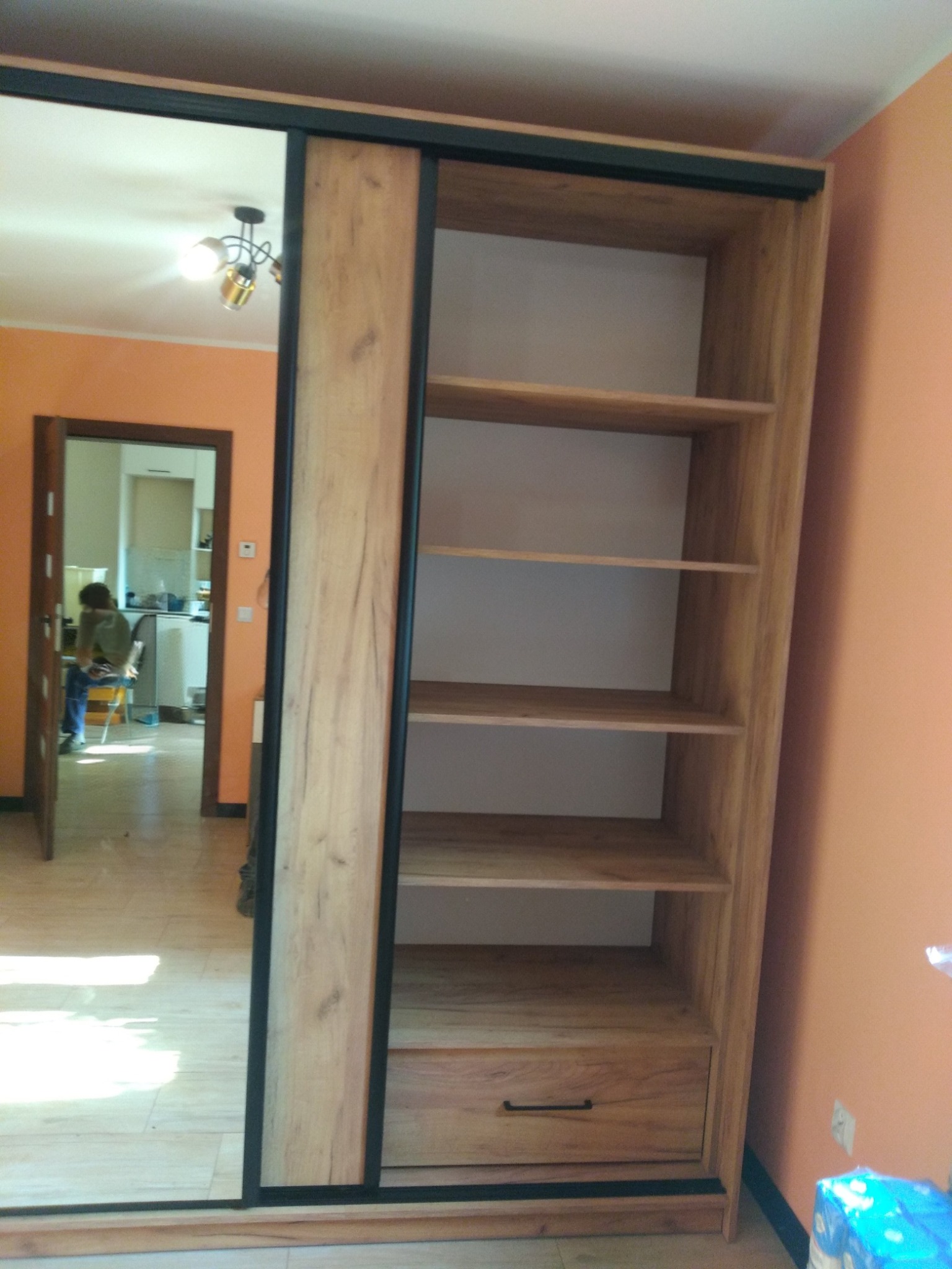 Szafa wnękowa z przesuwnymi drzwiami lustrzanymi i drewnianym wykończeniem, widoczne półki i szuflada. W tle pomarańczowa ściana i widok na kuchnię.