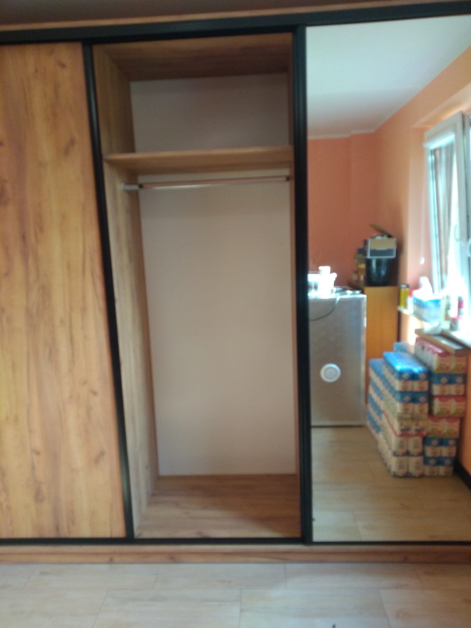 Szafa przesuwna z lustrem i drewnianymi frontami. Widoczne wnętrze szafy z półkami i drążkiem. W tle pokój z oknem i stertą kartonów.