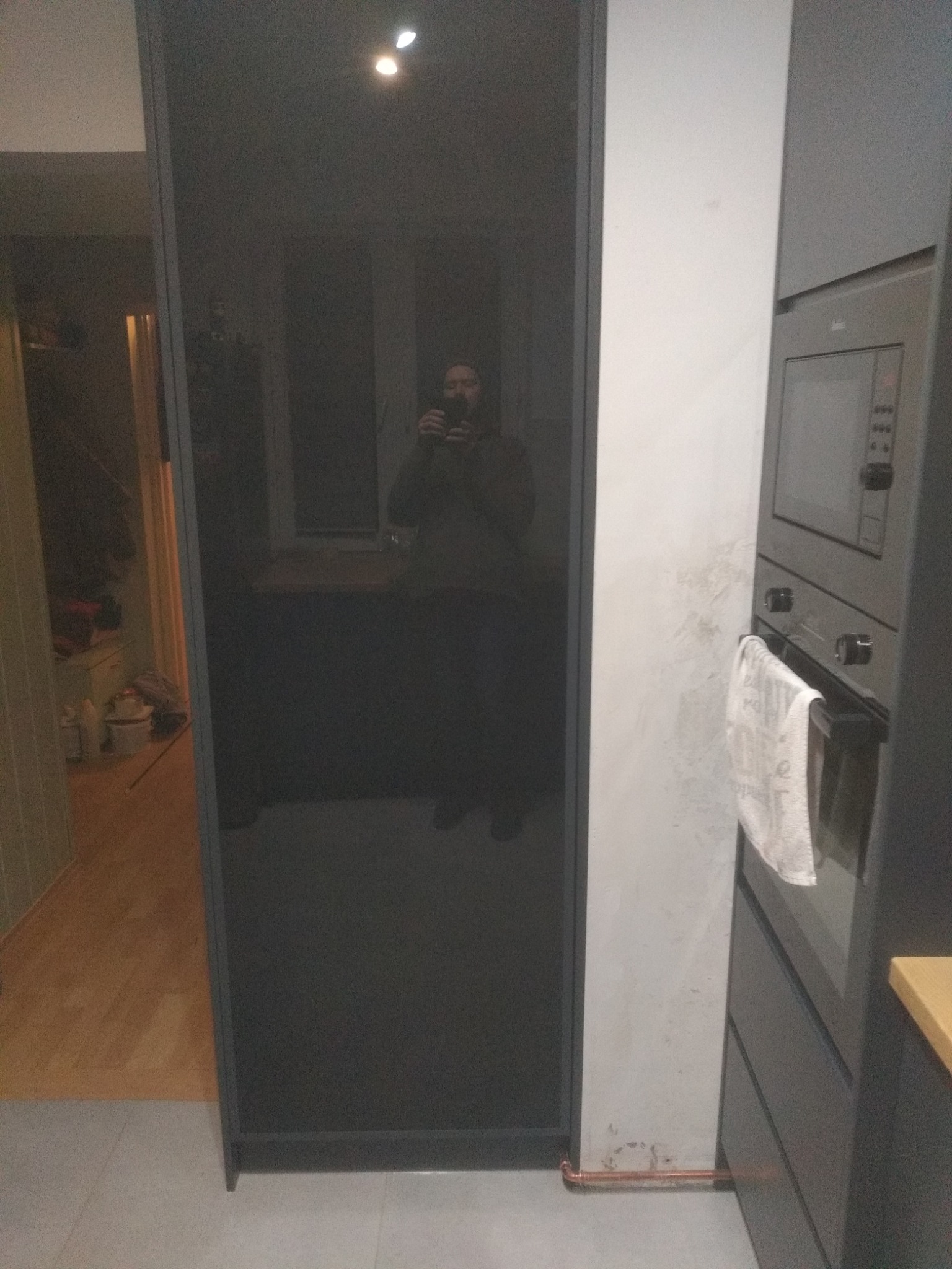 Ciemnoszara szafa wnękowa z lustrzanymi drzwiami, odbijająca wnętrze pokoju. Widoczny fragment zabudowy kuchennej z piekarnikiem i szarą szafką.