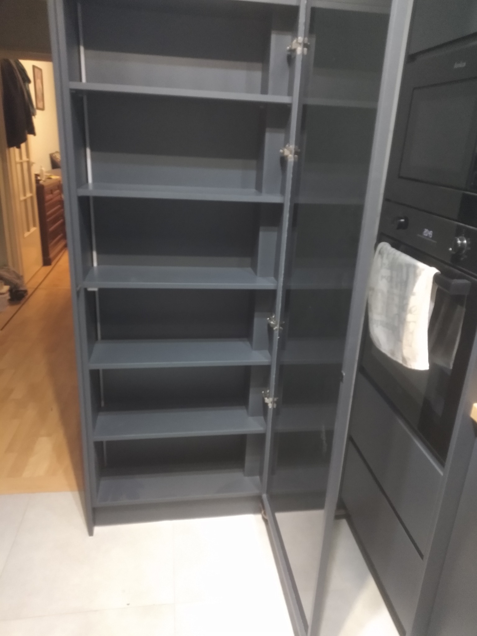 Szara szafa z otwartymi drzwiami w kuchni, widoczne półki. Obok zabudowany piekarnik. Ujęcie z bliska, widoczny fragment podłogi.