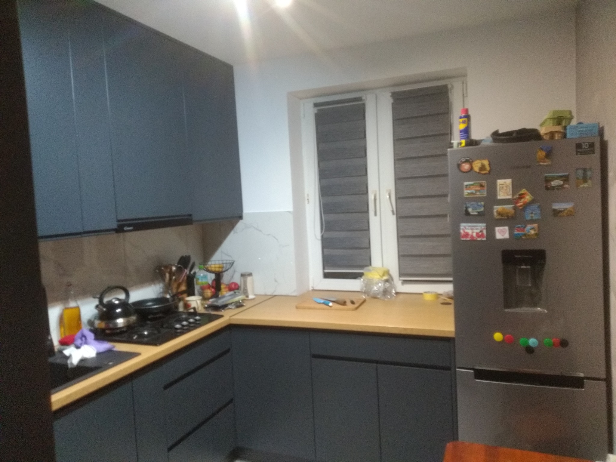 Nowoczesna kuchnia w odcieniach szarości z drewnianym blatem i marmurowym akcentem na ścianie, widok na okno z roletami i lodówkę z magnesami.