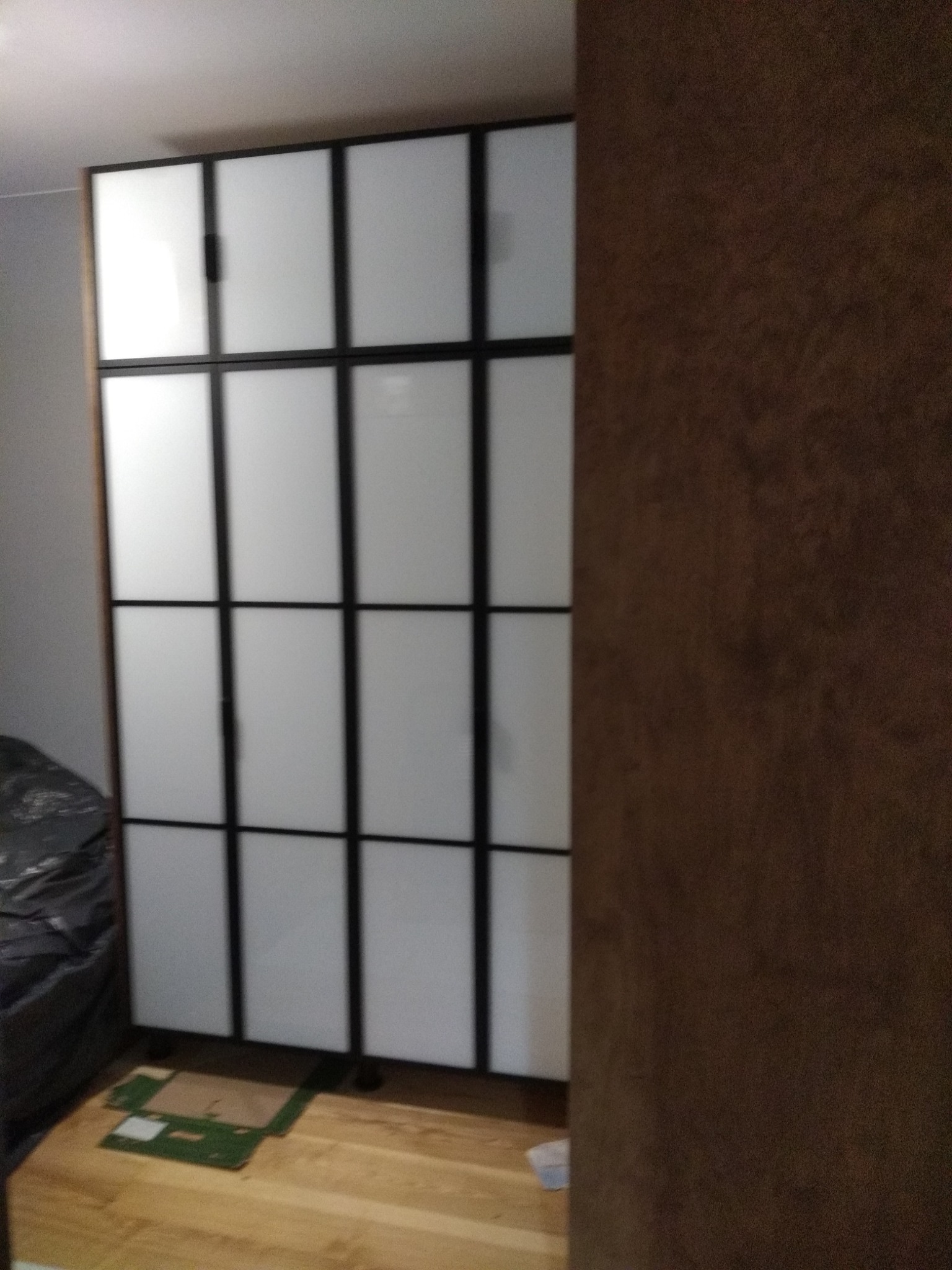 Szafa wnękowa z białymi frontami i czarnymi ramami, podzielona na kwadraty. Widoczna część ściany w kolorze brązowym i fragment podłogi.