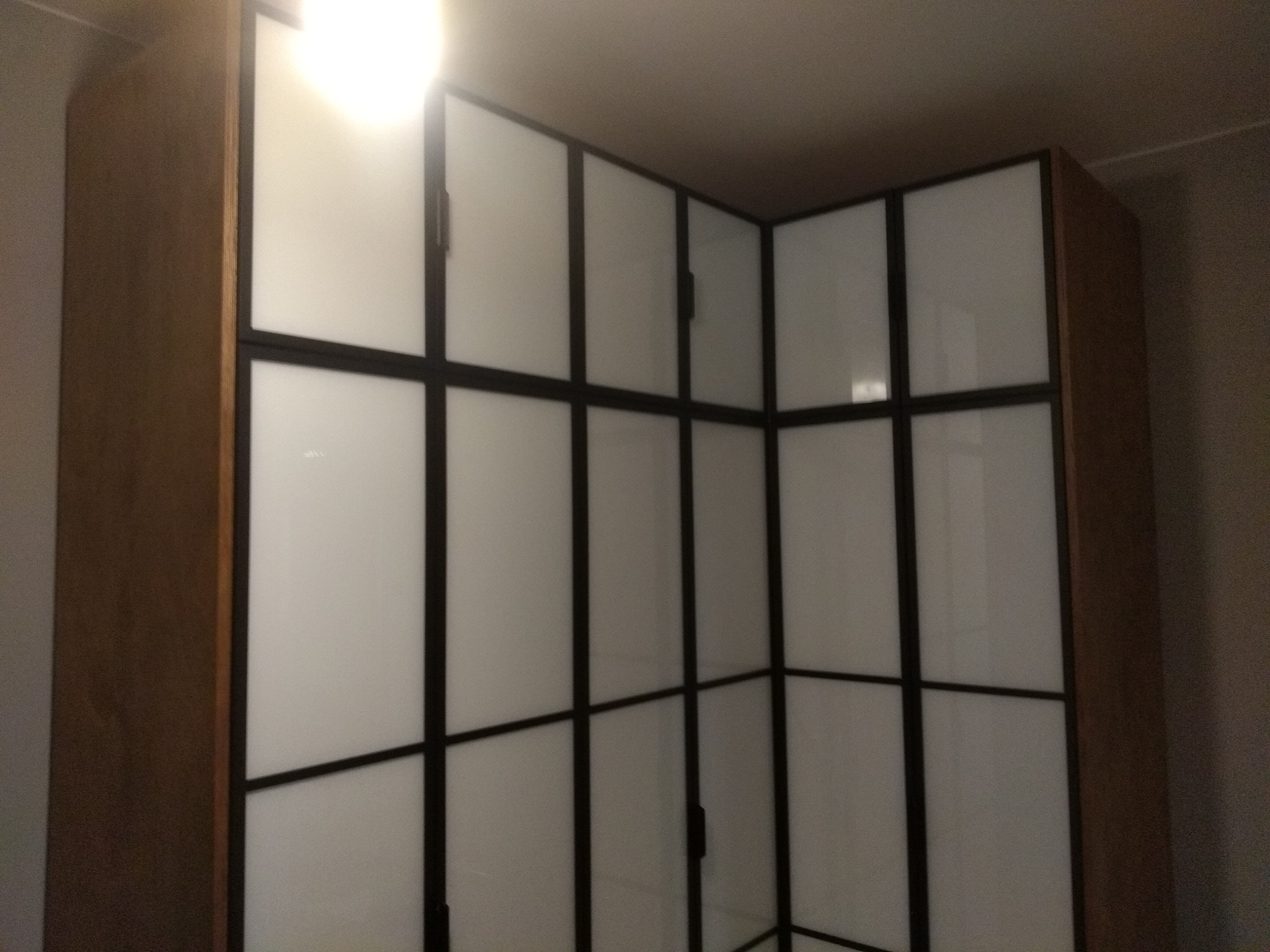 Narożna szafa z drewnianymi bokami i białymi frontami podzielonymi czarnymi listwami. Widoczne zawiasy i fragment sufitu. Ujęcie od dołu.