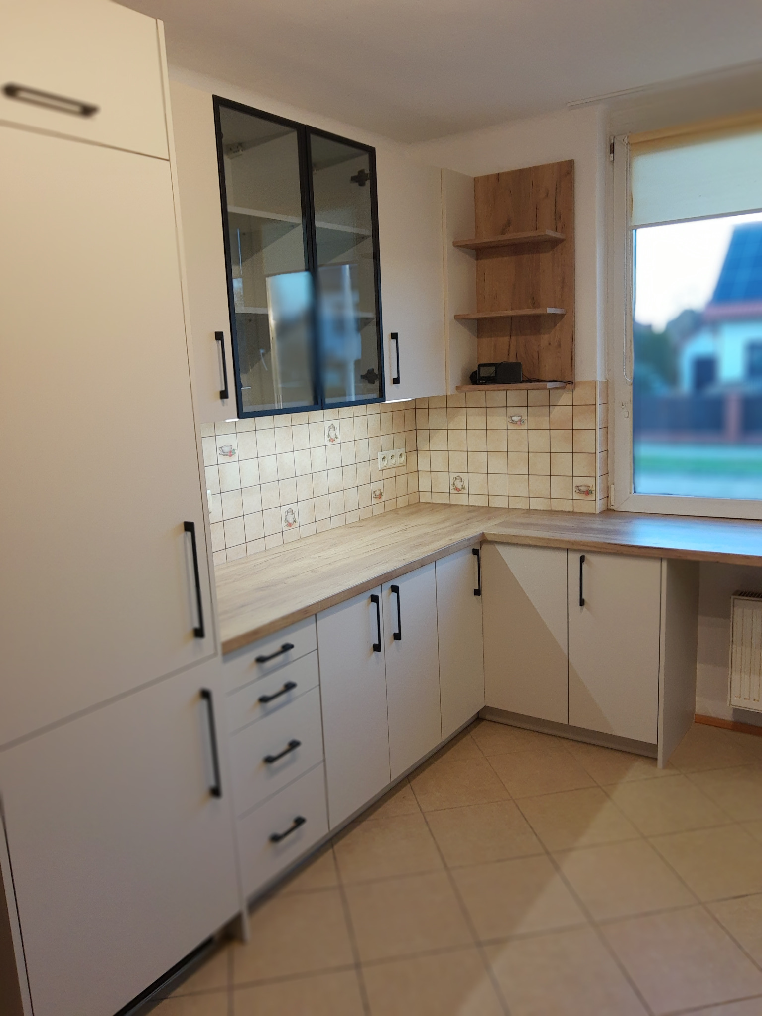 Jasna kuchnia z drewnianym blatem i czarnymi uchwytami. Na ścianie płytki z motywem filiżanek. Nad blatem szafka z przeszklonymi drzwiczkami i drewniane półki.