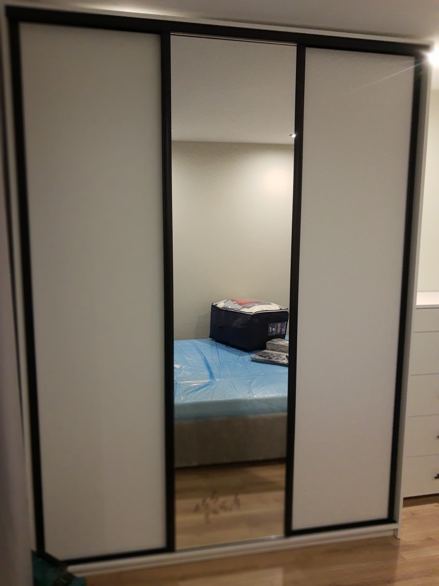 Trzydrzwiowa szafa przesuwna z lustrem w czarnej ramie. Widoczne odbicie łóżka z niebieskim prześcieradłem i komody. Minimalistyczny design, jasne wnętrze.