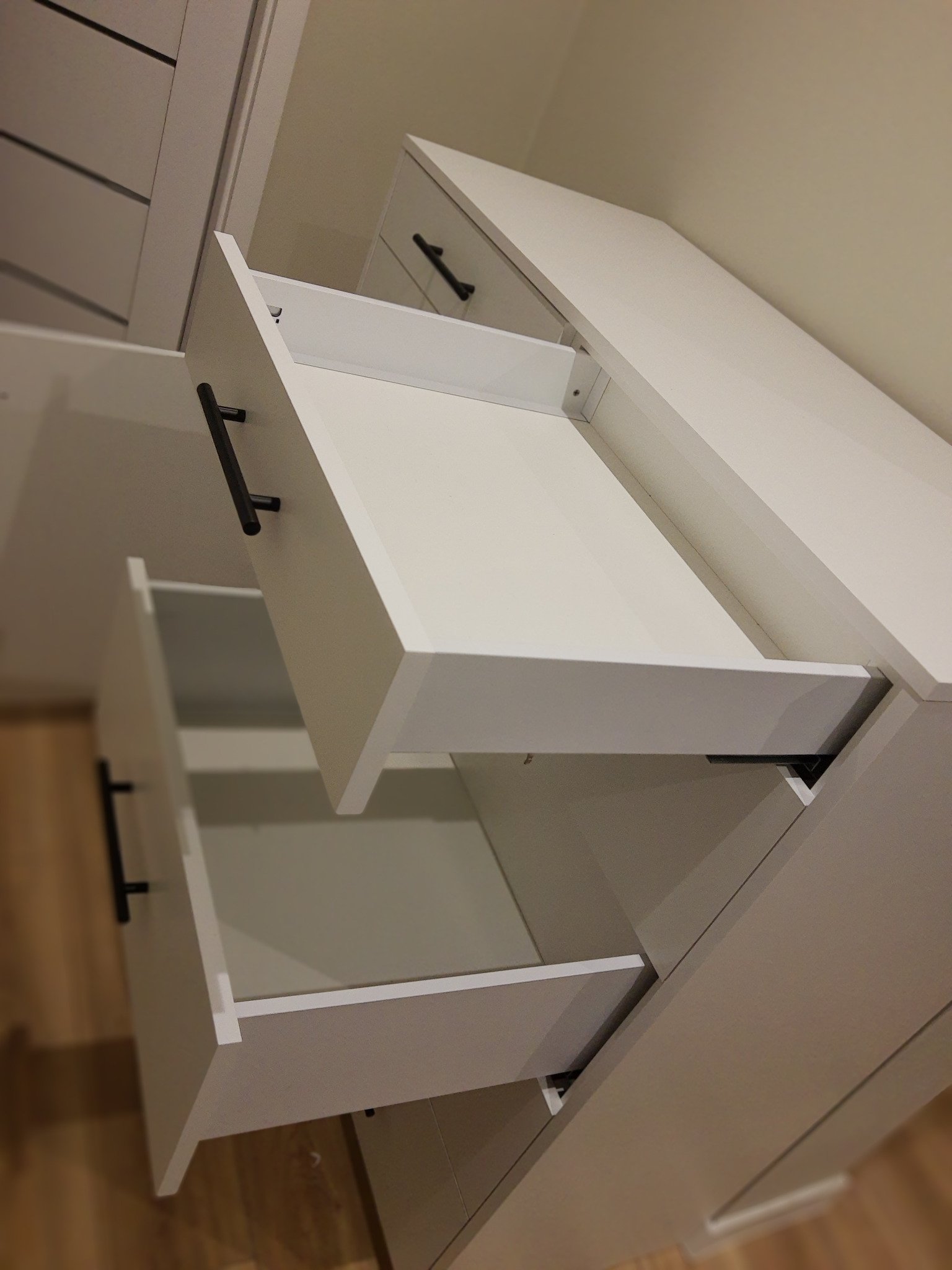 Biała komoda z wysuniętymi szufladami i czarnymi uchwytami, widok z dołu, ukazujący mechanizm wysuwania szuflad. Minimalistyczny design.