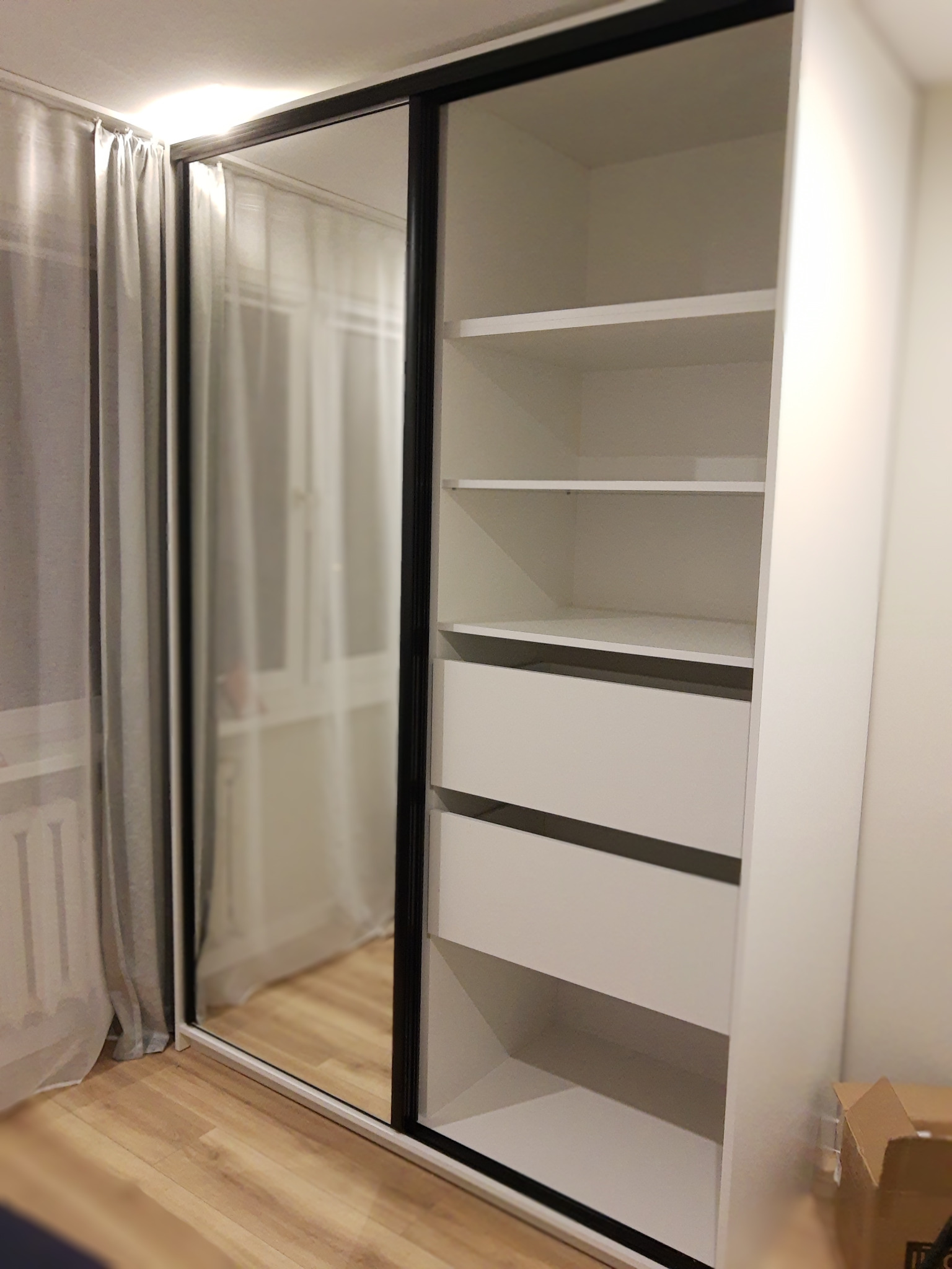 Biała szafa wnękowa z przesuwnymi drzwiami lustrzanymi i czarnymi obramowaniami, widoczne półki i szuflady, na podłodze panele, obok zasłona.