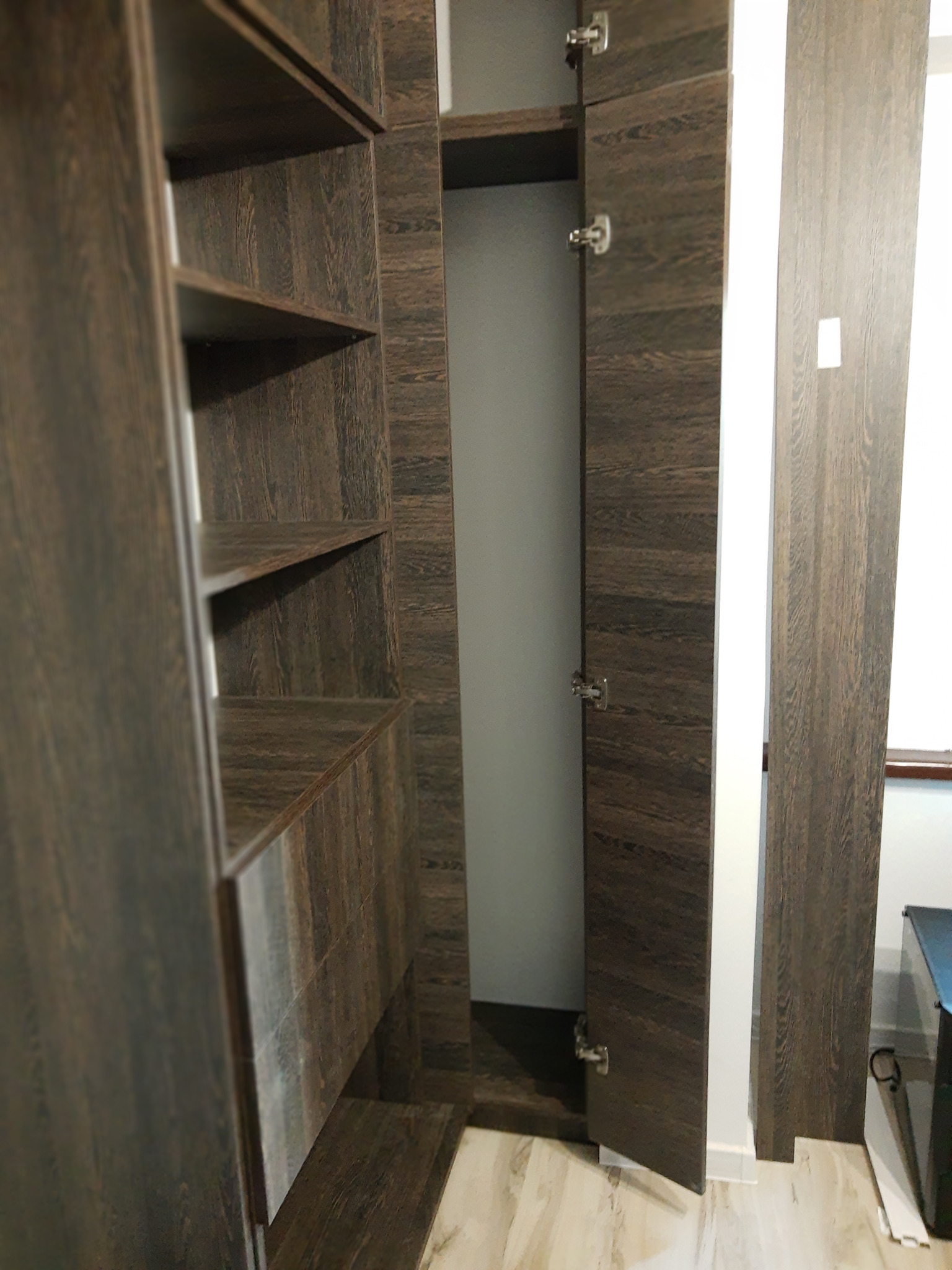 Wnętrze szafy wnękowej z otwartymi drzwiami, wykończonej w ciemnym drewnie z wyraźnym usłojeniem. Widoczne półki i system zawiasów. Ujęcie z bliska, podkreślające detale.