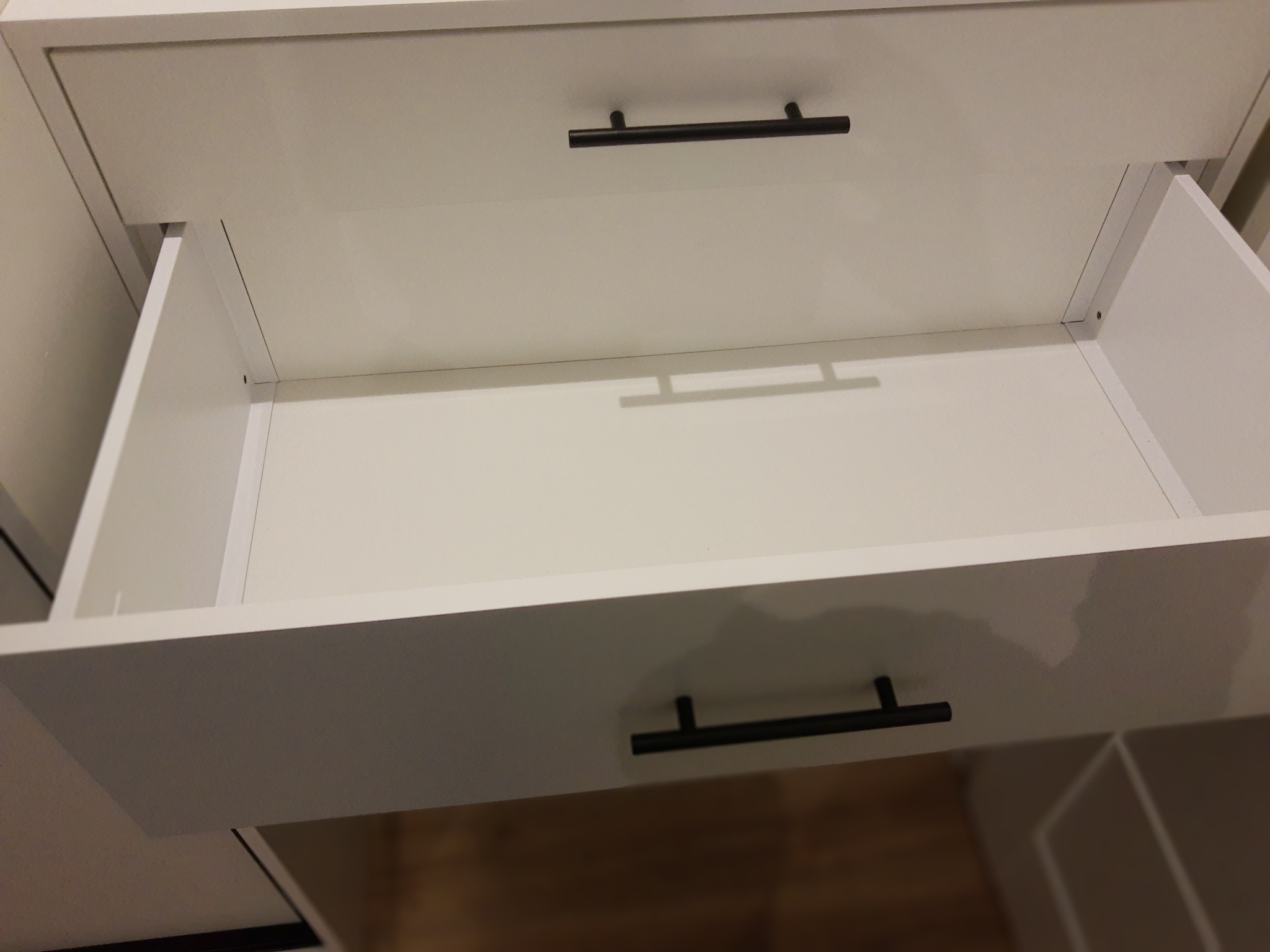 Otwarta, biała szuflada z czarnym, minimalistycznym uchwytem. Widoczna wewnętrzna konstrukcja szuflady i fragment szafki poniżej. Ujęcie z bliska, oświetlenie studyjne.