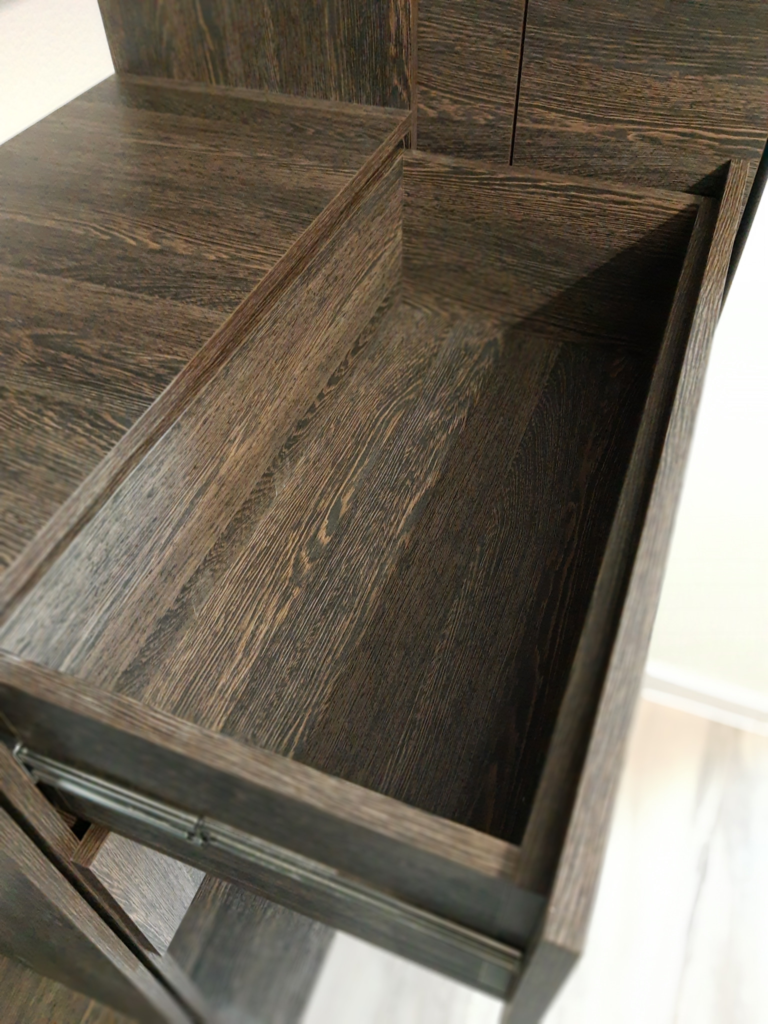 Wysunięta szuflada z ciemnego drewna z widoczną strukturą słojów, zamontowana na metalowych prowadnicach. Ujęcie z góry, ukazujące wnętrze szuflady i fragment mebla.