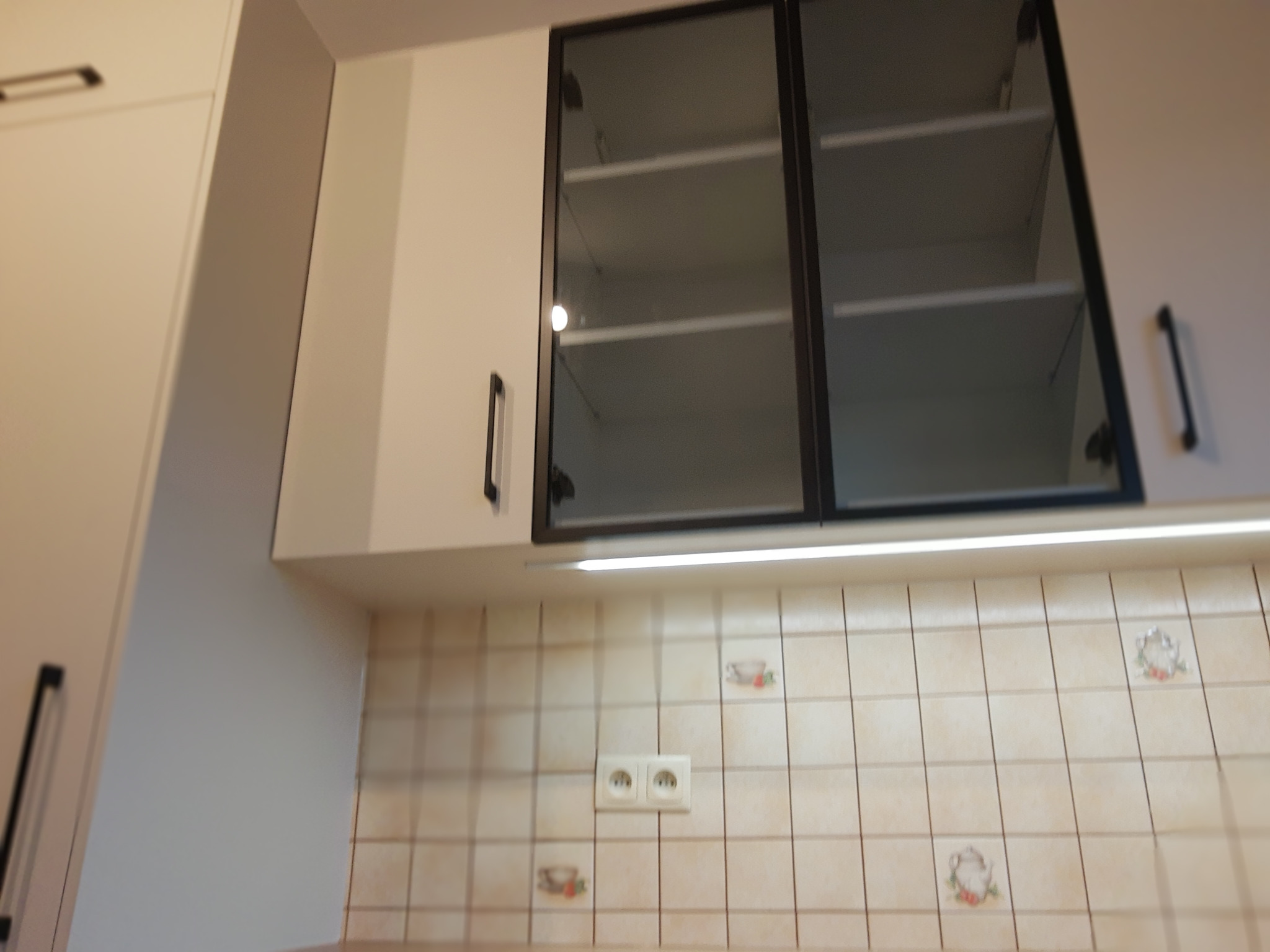 Białe szafki kuchenne wiszące z czarnymi uchwytami i szklanymi frontami, oświetlenie podszafkowe, kremowe płytki z motywem filiżanek w tle.