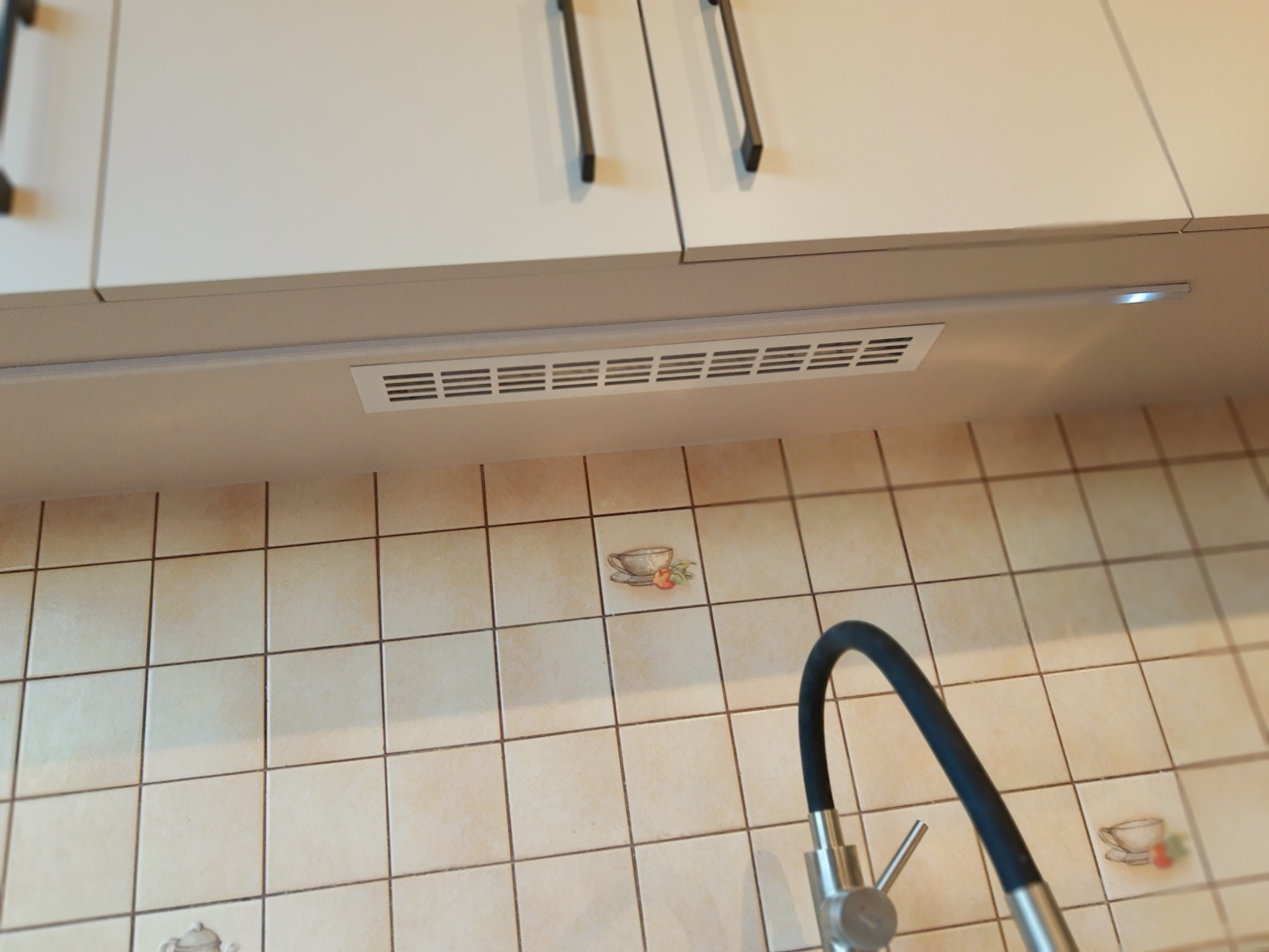 Białe szafki kuchenne z czarnymi uchwytami, oświetlenie podszafkowe LED i beżowe płytki z motywem filiżanki, widoczny fragment baterii kuchennej.