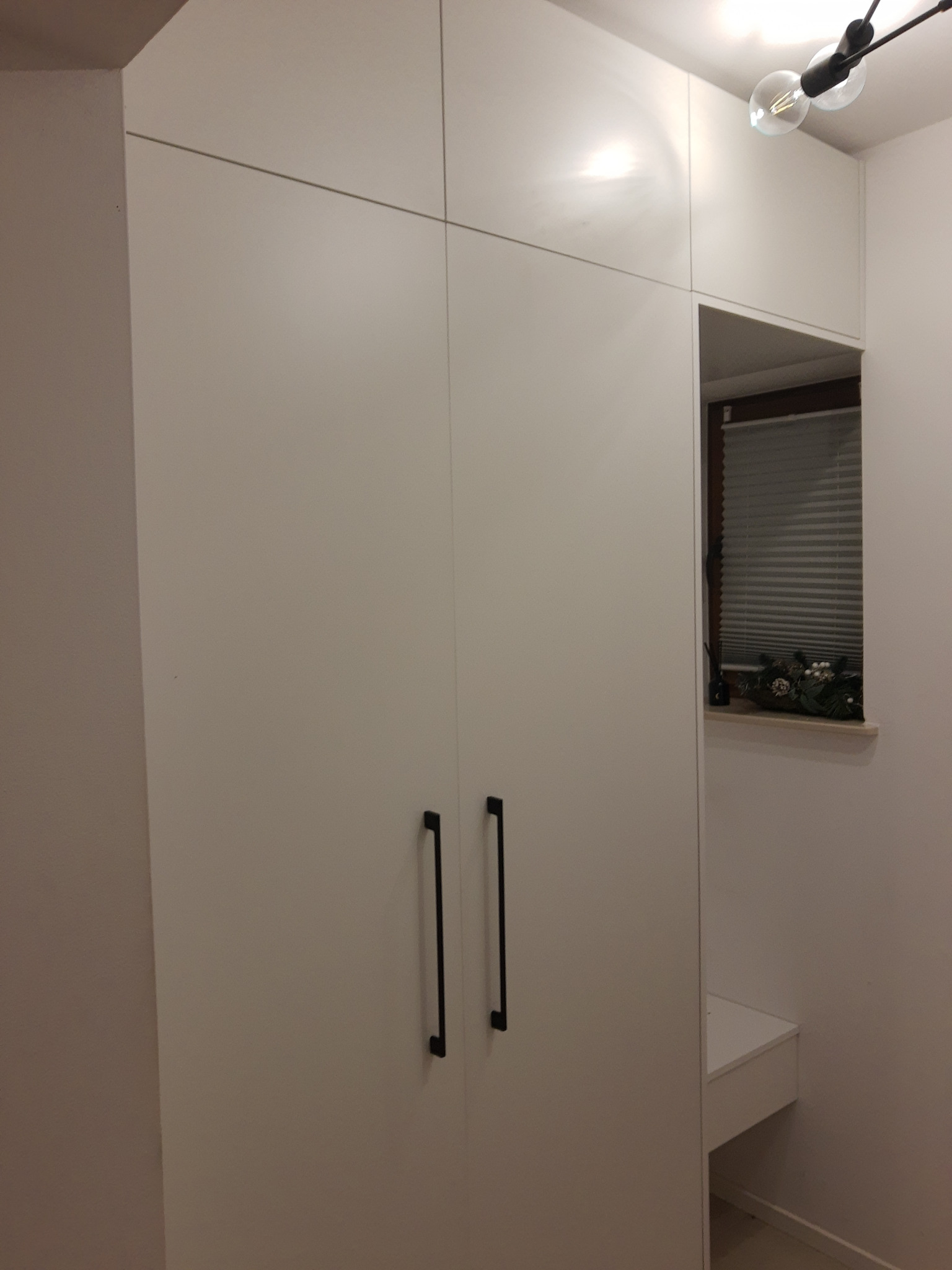 Biała szafa wnękowa z czarnymi uchwytami, wkomponowana we wnękę z oknem, minimalistyczny design, oświetlenie sufitowe w tle.