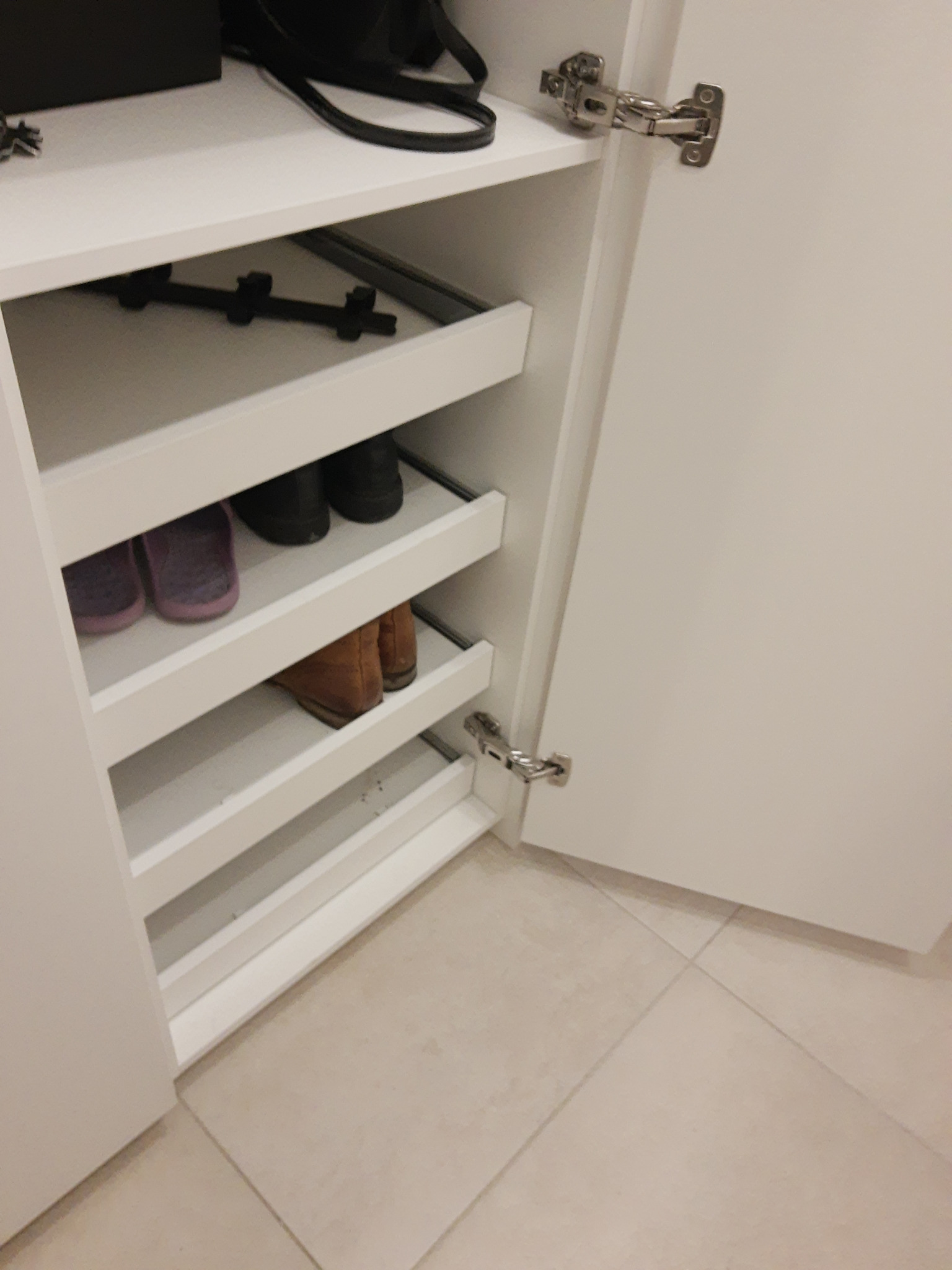 Wnętrze białej szafy z wysuwanymi półkami na buty, widoczne buty na półkach, otwarte drzwi szafy z zawiasami.