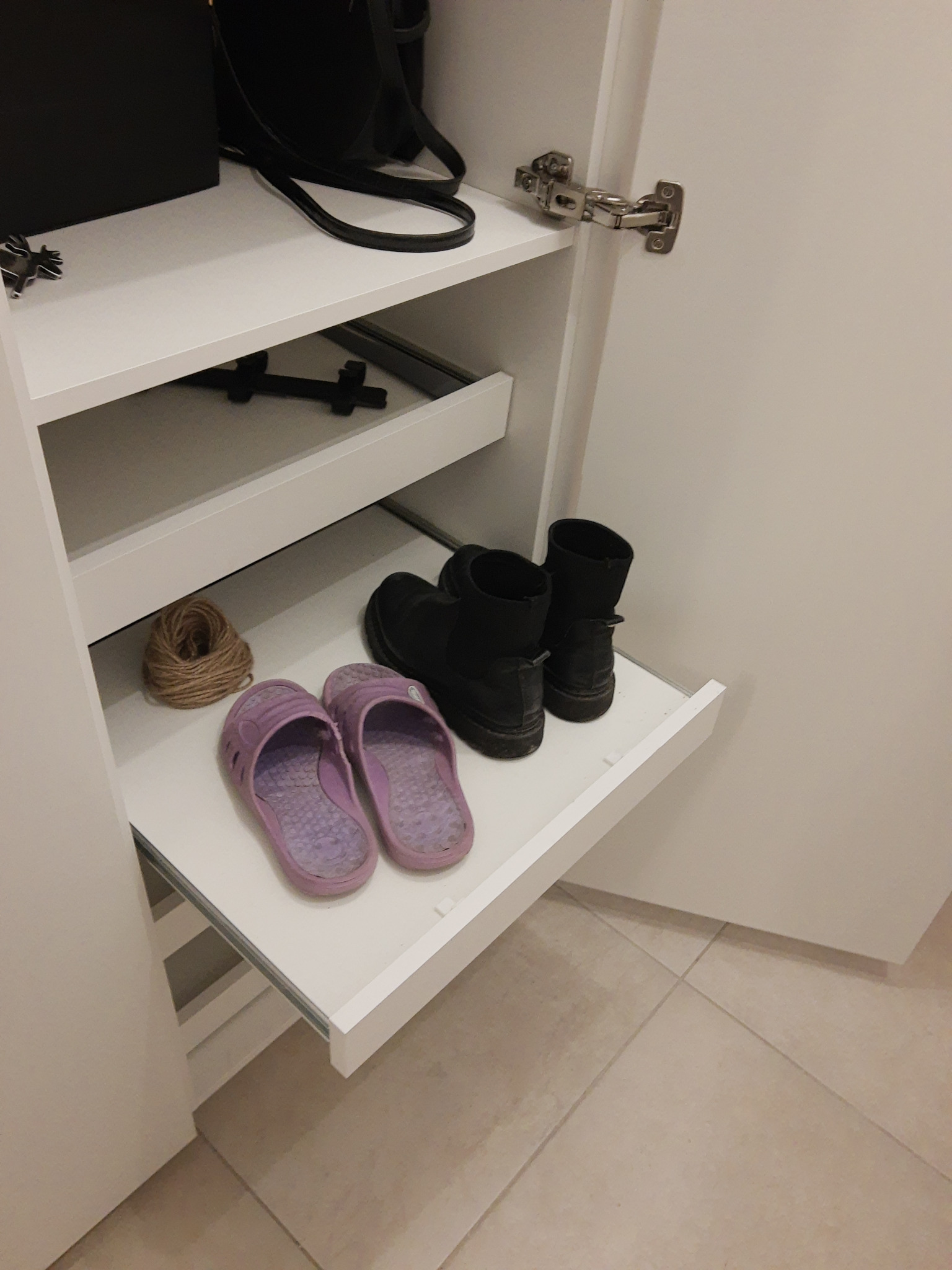 Wysuwana półka w szafie z zawartością: klapki, buty i sznurek. Widoczne okucia szafy i fragment podłogi z płytek. Ujęcie z bliska, wnętrze szafy w jasnej tonacji.