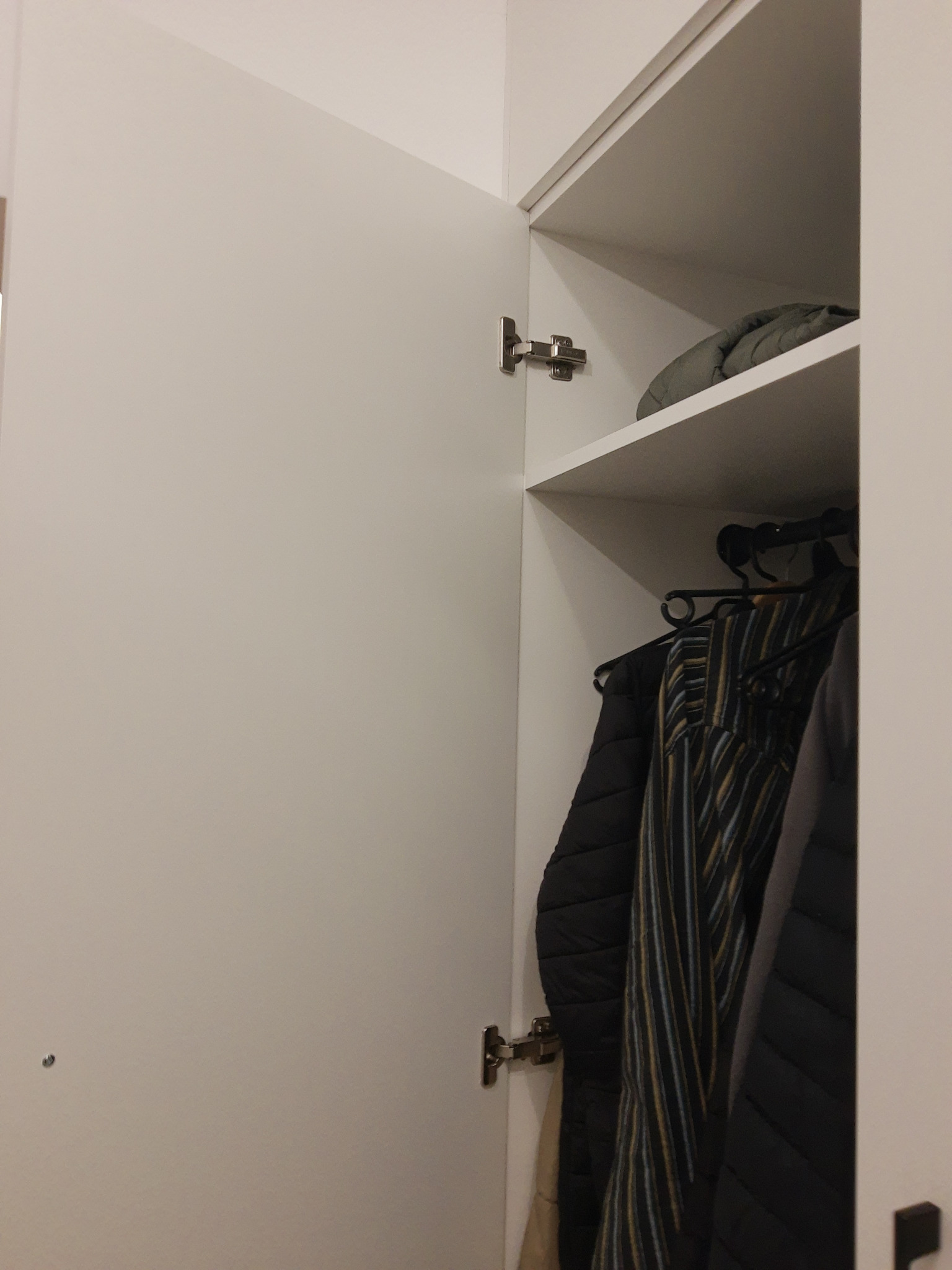 Otwarta, biała szafa wnękowa z widocznymi ubraniami na wieszakach i półką z kurtką. Metalowe zawiasy drzwi szafy. Minimalistyczny design.