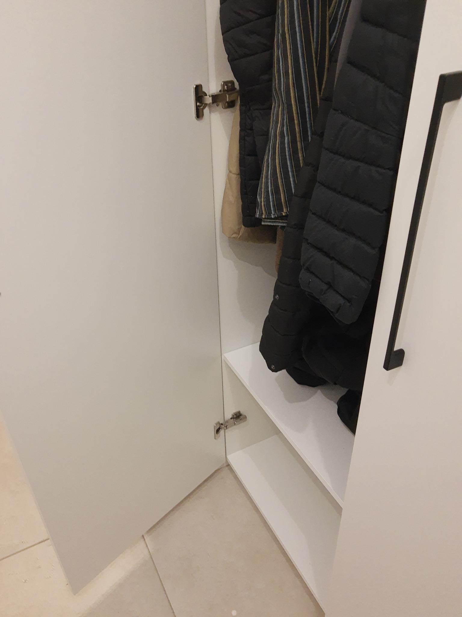 Otwarta, biała szafa wnękowa z ubraniami w środku, widoczne półki i zawiasy. Minimalistyczny design, jasne wnętrze.