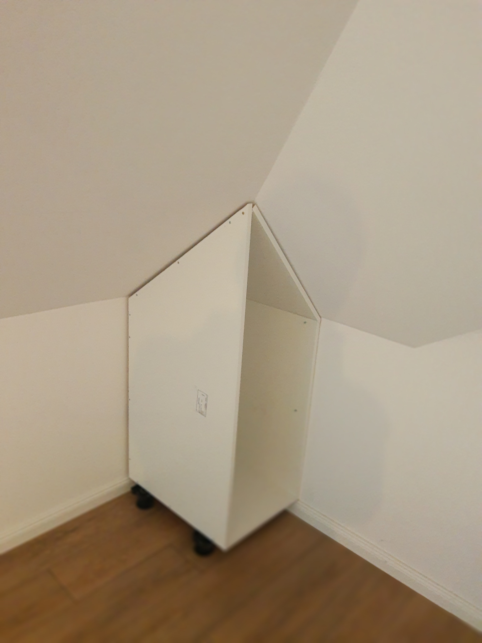 Biała szafka na kółkach, dopasowana do skosu poddasza, stoi przy ścianie na drewnianej podłodze. Widoczne łączenia płyt i śruby.