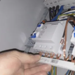 Dłoń elektryka przy listwie zaciskowej w rozdzielnicy elektrycznej, widoczne przewody o różnych kolorach.