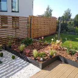 Ogród z podwyższonymi rabatami, wysypany korą i żwirem, otoczony drewnianym płotem i drewnianym tarasem, z widocznymi różnymi roślinami w donicach i bez