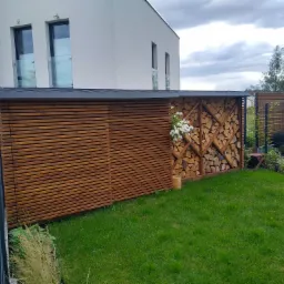 Drewniana konstrukcja z poziomych listew, częściowo ażurowa z ułożonym drewnem, z białymi kwiatami, na tle zielonego trawnika i nowoczesnego domu.