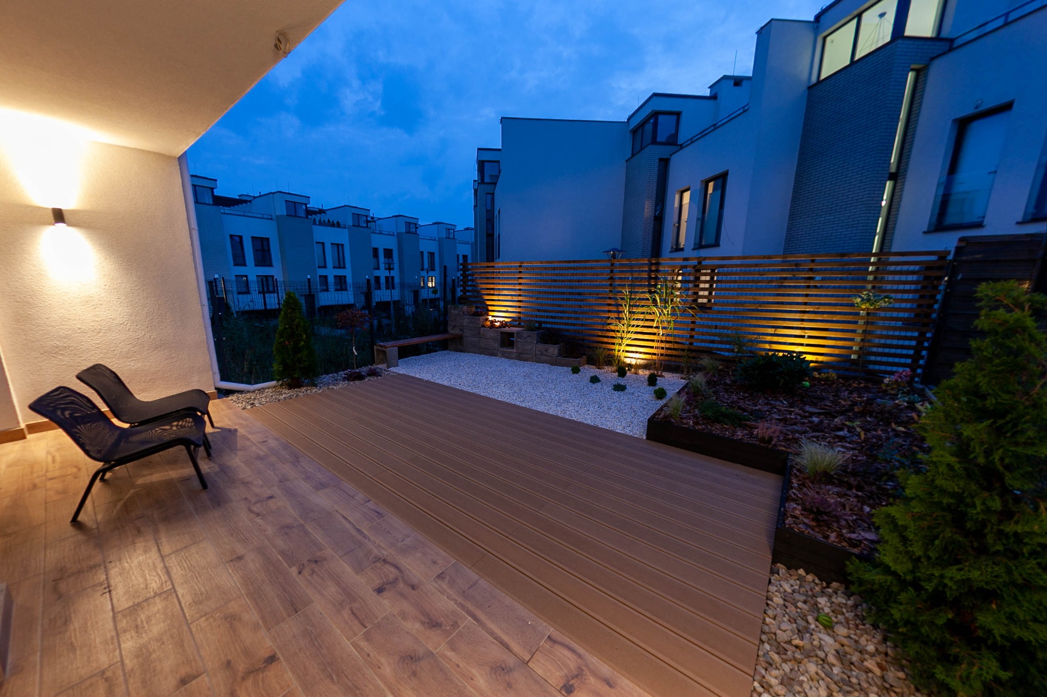 Nowoczesny taras z desek kompozytowych, oświetlony punktowo, z minimalistycznym ogrodem żwirowym i drewnianą ażurową ścianką, widok na szereg domów w tle, wieczorne oświetlenie.