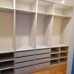 Przestronna, biała garderoba z szufladami i półkami, wykonana na wymiar, z widoczną strukturą drewna i oświetleniem punktowym.