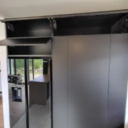 Szara szafa wnękowa z otwieranymi do góry szafkami, widoczne zawiasy i wnętrze szafek, w lustrzanych drzwiach odbija się warsztat stolarski.