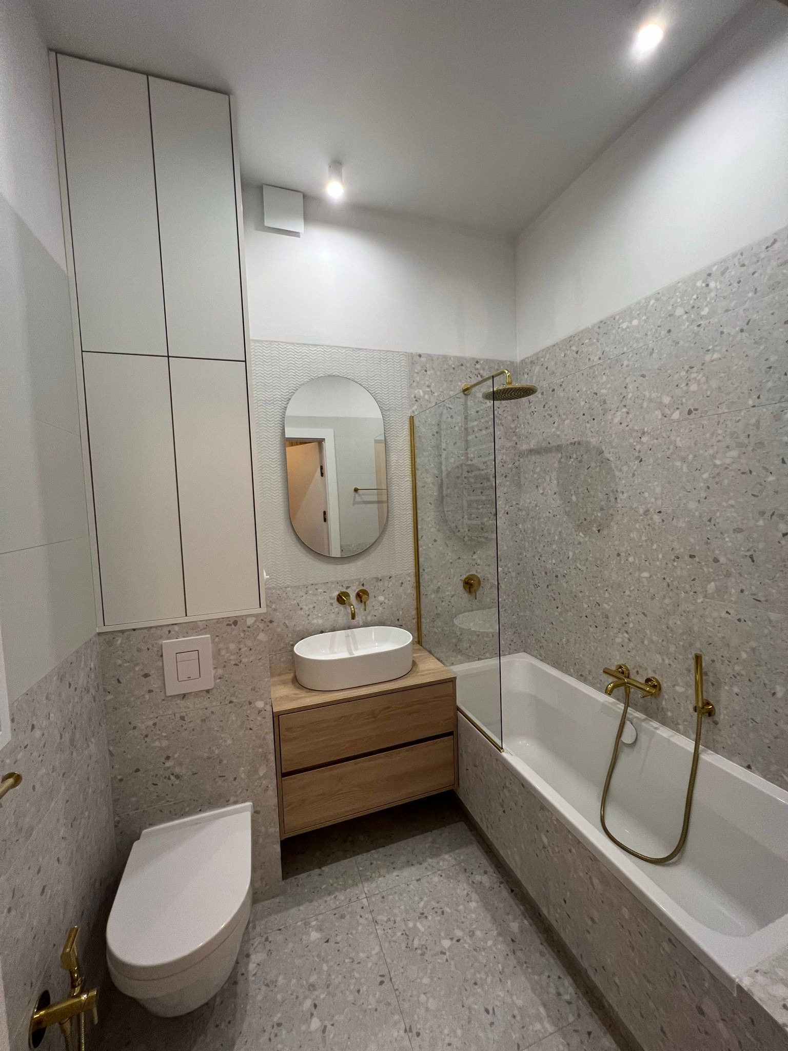 Nowoczesna łazienka z szarymi płytkami w stylu lastryko, złotą armaturą i owalnym lustrem.