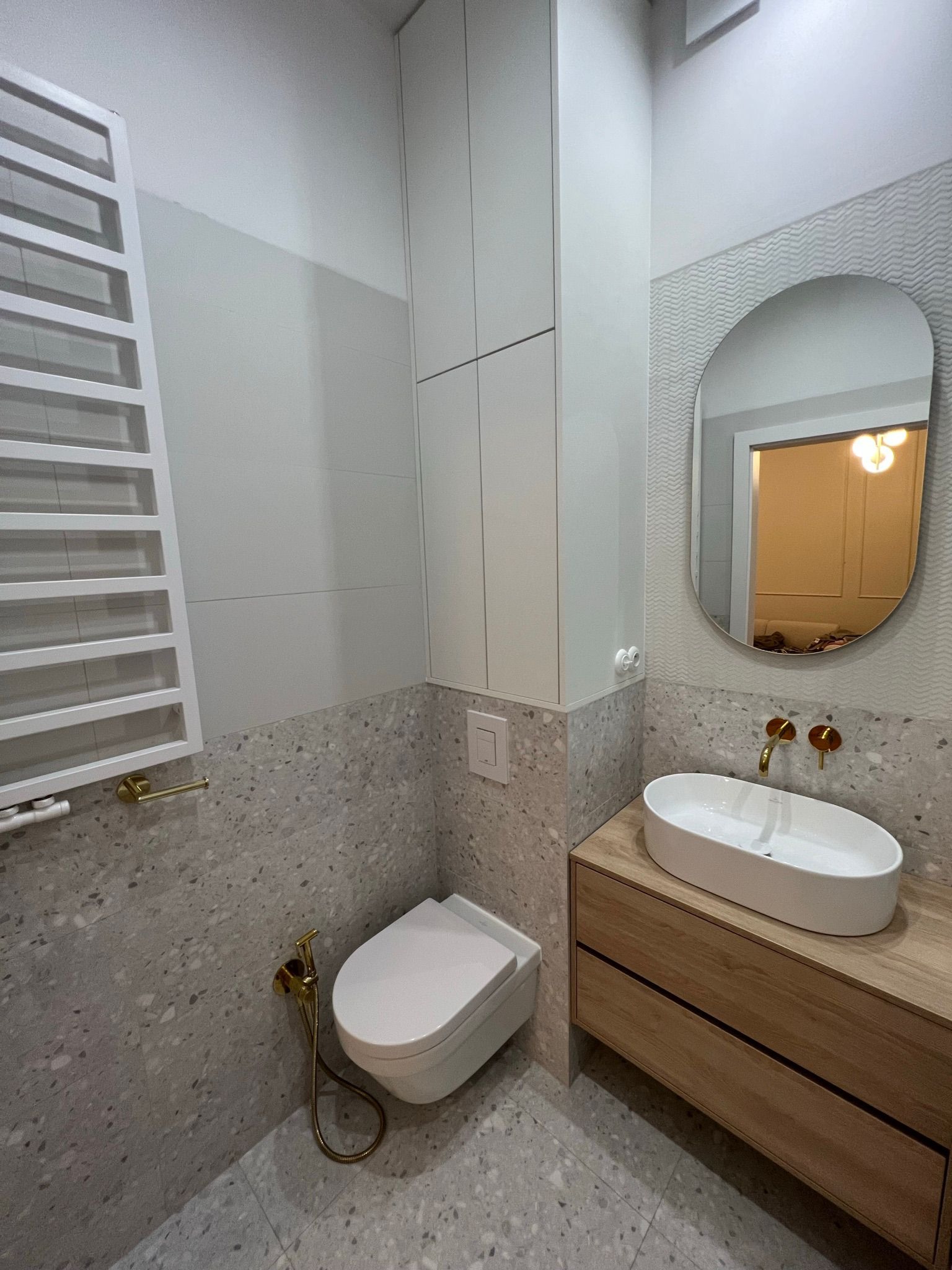 Nowoczesna łazienka z terazzo, wisząca miska WC z bidetką w kolorze złotym, umywalka nablatowa na drewnianej szafce, owalne lustro.