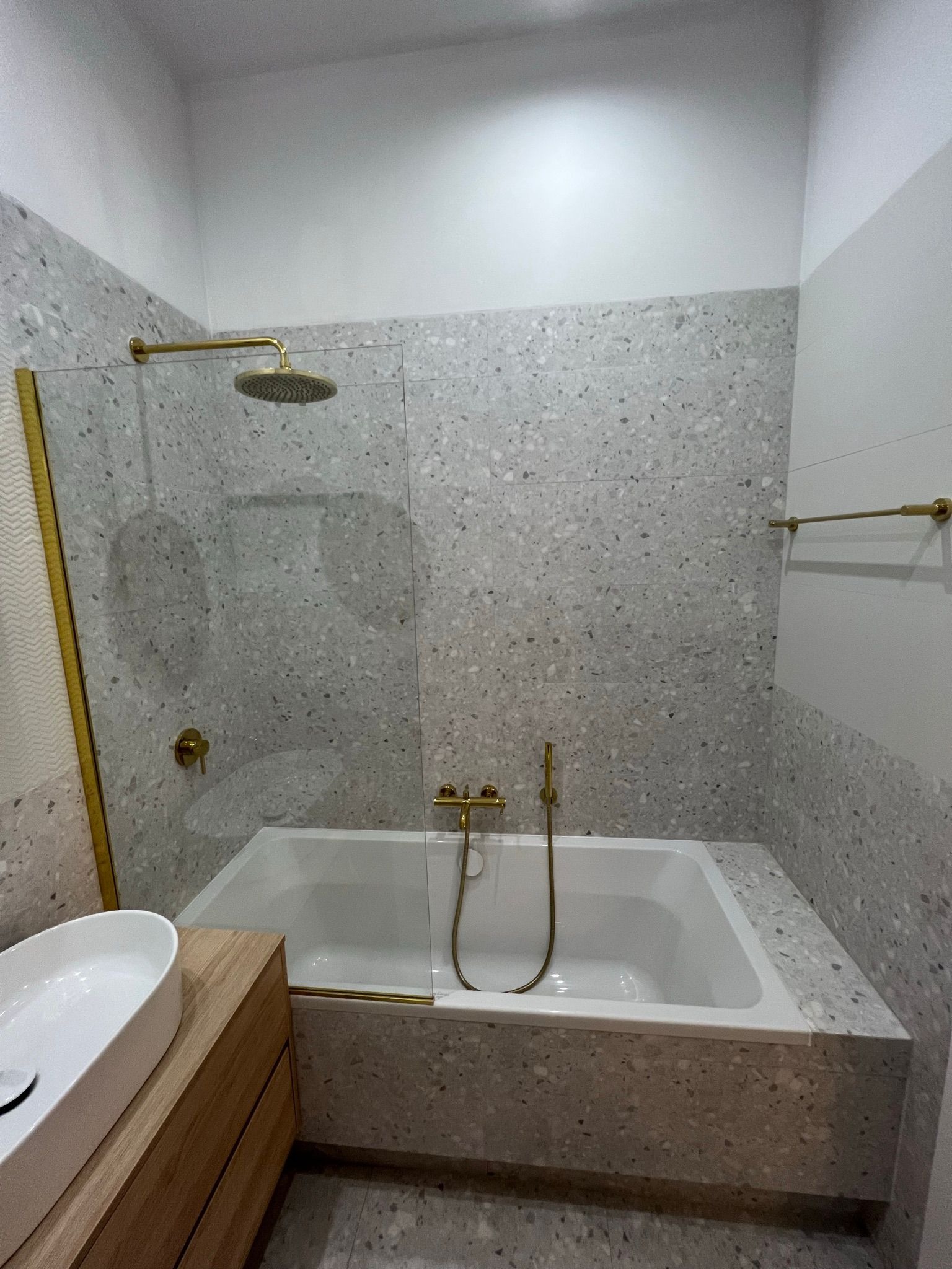 Wykończona łazienka w Warszawie z wanną obudowaną płytkami lastryko, złotą armaturą i szklanym parawanem nawannowym.