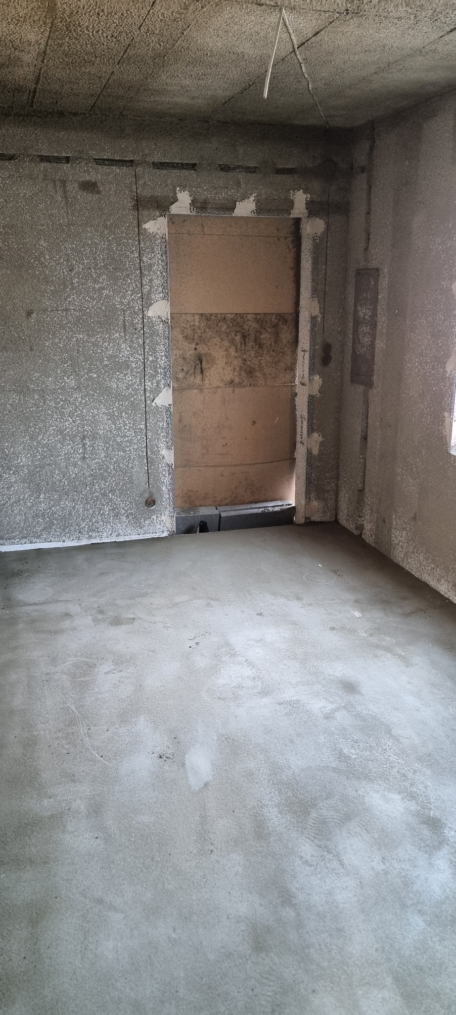 Surowe wnętrze w trakcie prac wykończeniowych: świeżo wylana posadzka, ściany z szorstkim tynkiem, zabezpieczone otwory drzwiowe.