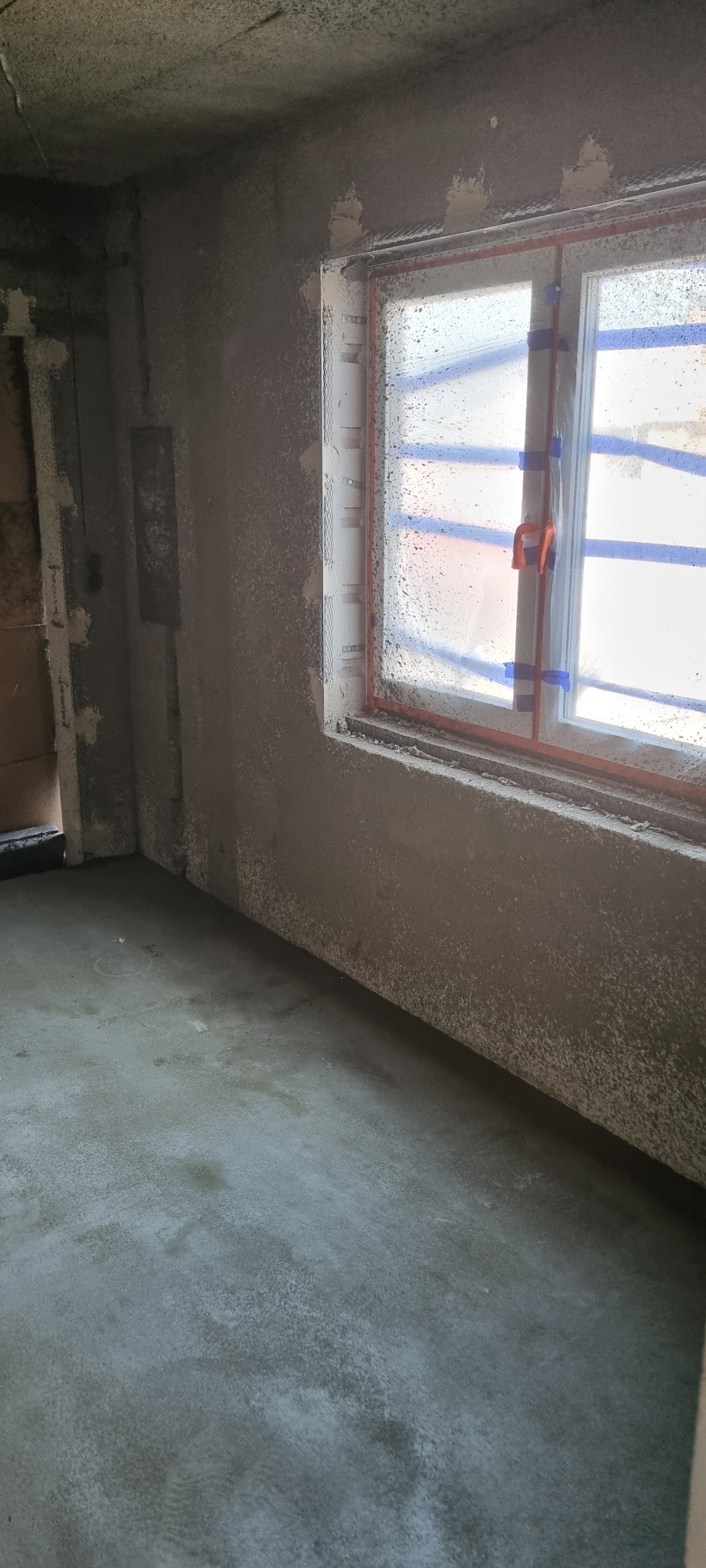 Świeżo nałożony, szary tynk maszynowy na ścianach i podłodze w pomieszczeniu z oknem zabezpieczonym folią i taśmą malarską.