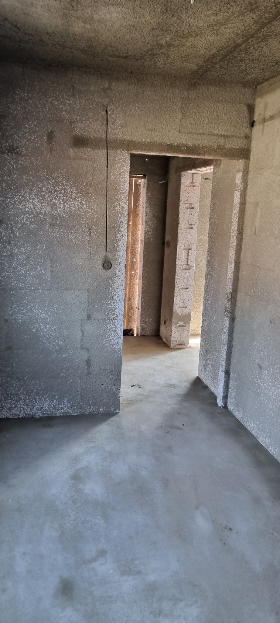 Surowe wnętrze z widoczną strukturą tynku na ścianach i suficie, zarys instalacji elektrycznej, widok na kolejne pomieszczenia w stanie deweloperskim.