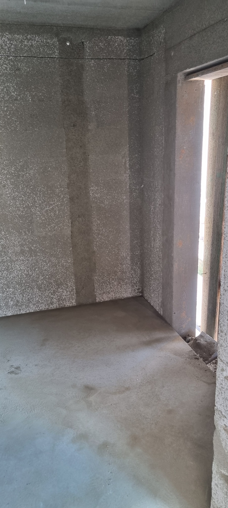 Wnętrze pomieszczenia po nałożeniu szarego tynku maszynowego na ściany i podłogę, widoczne pionowe pasy wzmocnienia na ścianach oraz surowe obramowanie drzwi z widocznymi odłamkami gruzu.