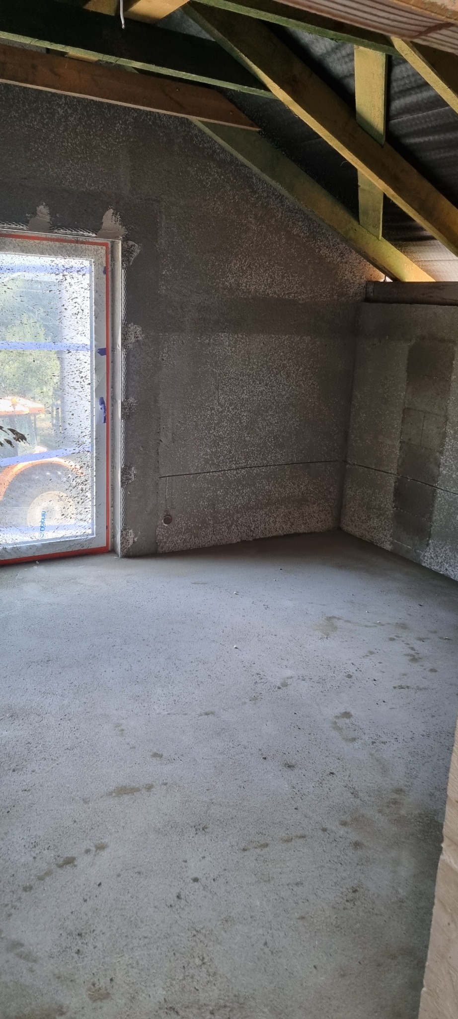 Wnętrze poddasza po nałożeniu tynków maszynowych, widoczna surowa podłoga, ściany pokryte świeżym tynkiem oraz drewniana konstrukcja dachu. Okno z widocznym na zewnątrz traktorem, zasłonięte...